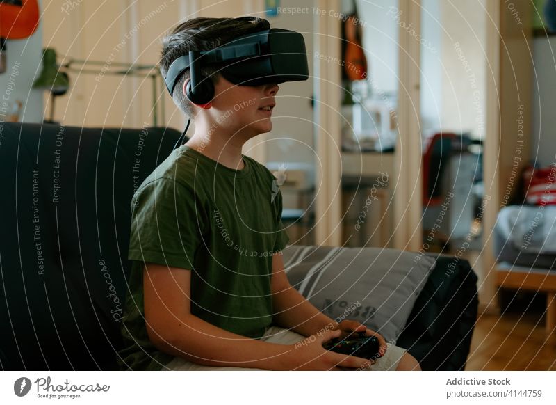 Kind in VR-Headset spielt auf der Couch Junge Schutzbrille spielen Gamepad virtuell Realität benutzend Sofa unterhalten modern heimwärts Gerät wenig bezaubernd