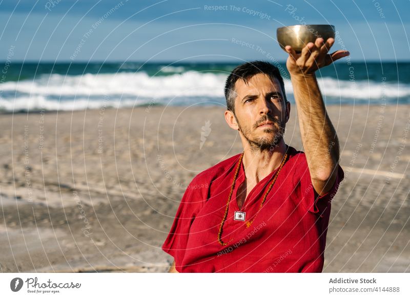 Friedlicher Mann benutzt tibetische Klangschale zum Stressabbau am Strand spielen Meer meditieren Tradition Kultur sich[Akk] entspannen wolkenlos Blauer Himmel