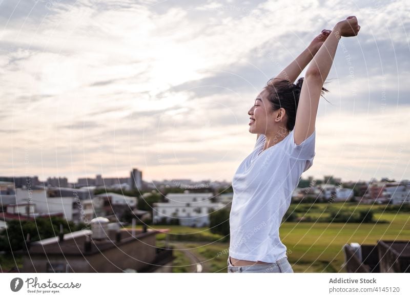 Glückliche asiatische Frau streckt ihren Körper am Morgen Dehnung Lächeln Arme hochgezogen Himmel wolkig Sonnenaufgang Terrasse jung ethnisch Freude