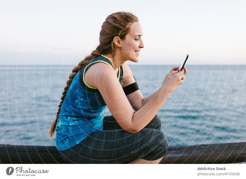 Lächelnde Frau in Sportkleidung mit Smartphone am Meer Sportlerin Stauanlage Browsen sich[Akk] entspannen Training MEER Sportbekleidung benutzend Fitness