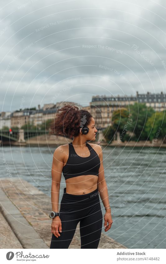 Ethnische Sportlerin mit Headset beim Training in der Nähe des Stadtflusses Athlet zuhören Musik intelligente Uhr Energie Fluss benutzend Gerät Apparatur