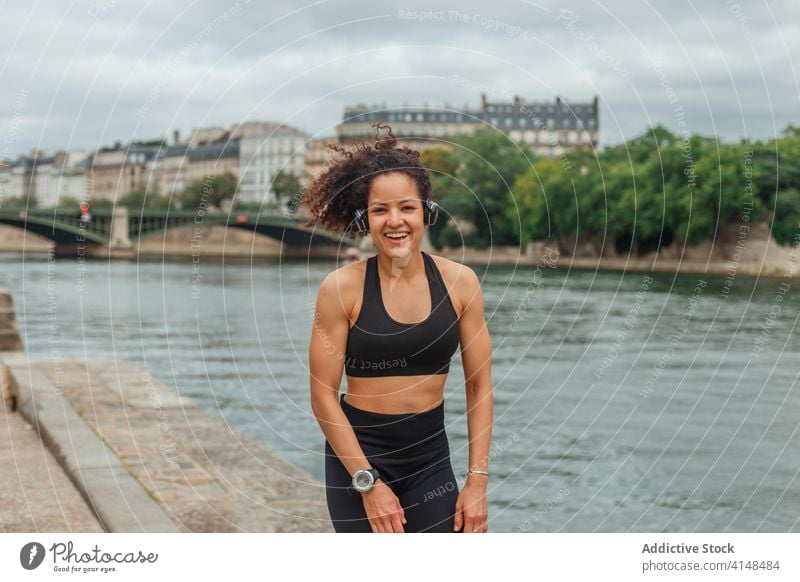 Fröhliche ethnische Sportlerin beim Headset-Training am Stadtfluss Athlet zuhören Musik intelligente Uhr Energie Fluss benutzend Gerät Apparatur Großstadt