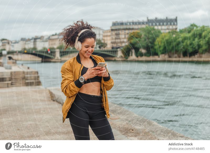 Sportlerin mit Headset nutzt Social Media auf Smartphone nach dem Training Kopfhörer soziale Netzwerke plaudernd Internet Pause Stauanlage Fluss benutzend