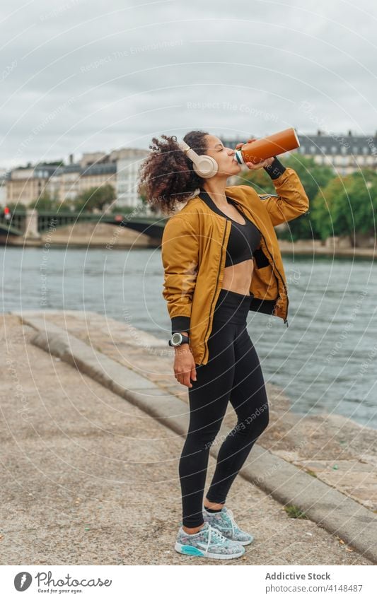 Durstige ethnische Sportlerin trinkt Wasser, nachdem sie in der Nähe eines Flusses trainiert hat trinken Flasche Headset Sportbekleidung Pause Baum benutzend