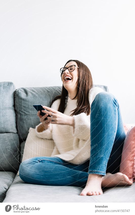 Frau, die ein Mobiltelefon benutzt, während sie zu Hause auf einer Couch sitzt. im Innenbereich benutzend arbeiten Arbeit Büro Gespräch Job Notebook jung Glück