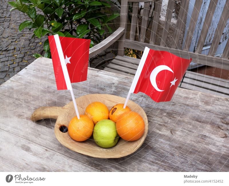 Orangen mit einem grünen Apfel und roten türkischen Wimpeln in einer Holzschale auf einem alten Holztisch mit Holzbank in den Gassen der Altstadt von Alacati bei Izmir in der Türkei