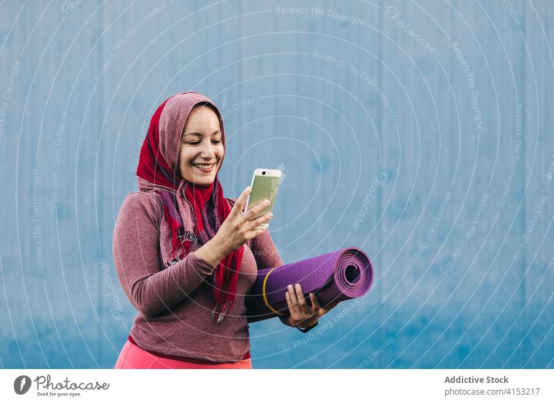 Lächelnde ethnische Sportlerin mit Smartphone in der Stadt benutzend Browsen Frau Hijab muslimisch Unterlage arabisch Großstadt Training Telefon Mobile Straße