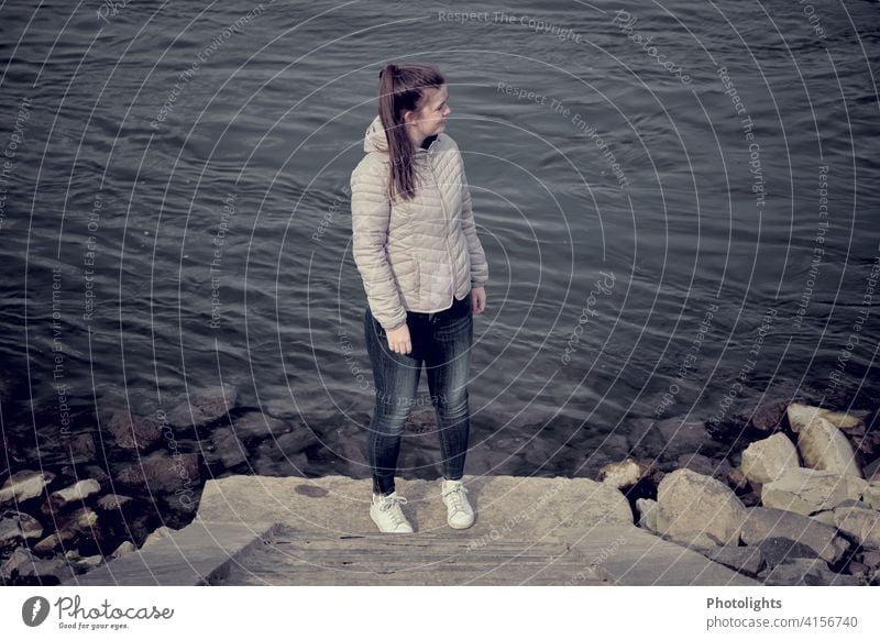 Junge Frau steht am Ufer eines Flusses jung Steine Steintreppe Wasser dunkel düster alleine Natur Außenaufnahme Farbfoto Landschaft Pferdeschwanz einsam