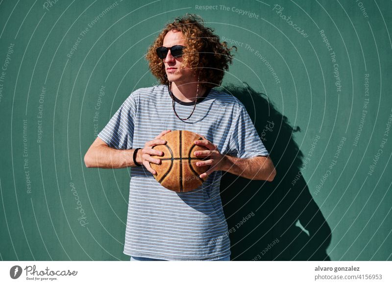 ein junger Mann mit einem Basketball und Sonnenbrille mit einer grünen Wand im Rücken, der in die Sonne schaut che Ball Person Korb Sport sportlich männlich