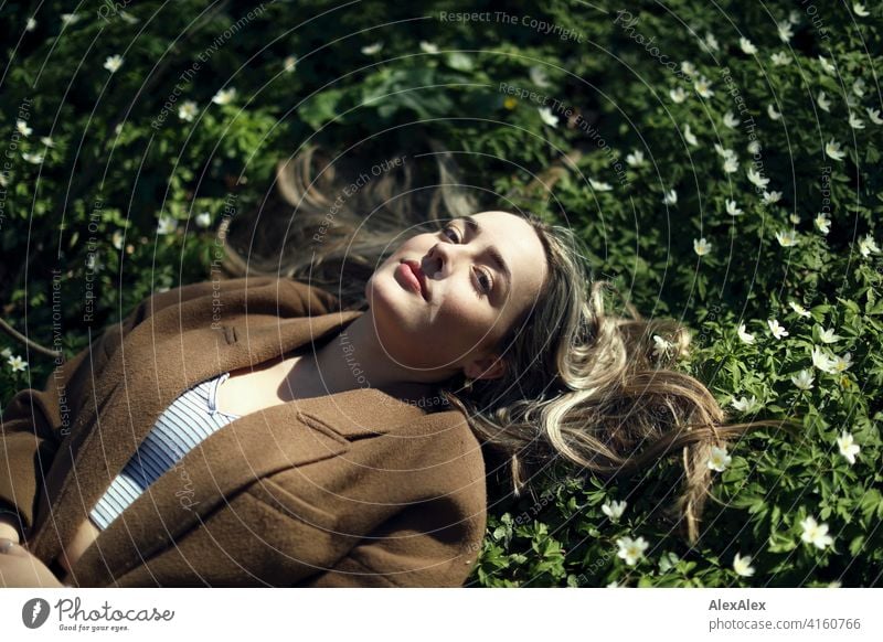 Ein blondes Mädchen liegt im Wald in einer blühenden Wiese auf der Lichtung mit kleinen weißen Blumen und schaut nach oben in die Kamera Landschaft Freude