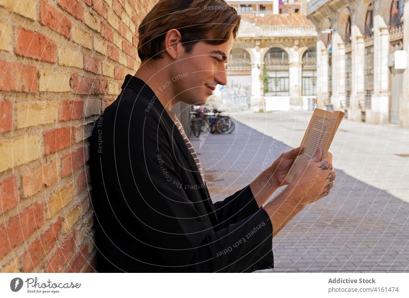 Crop lächelnd trendy tätowierten Mann lesen Buch auf dem Bürgersteig Vorschein Literatur Lächeln Tattoo Backsteinwand Straßenbelag Stadt stylisch Bekleidung