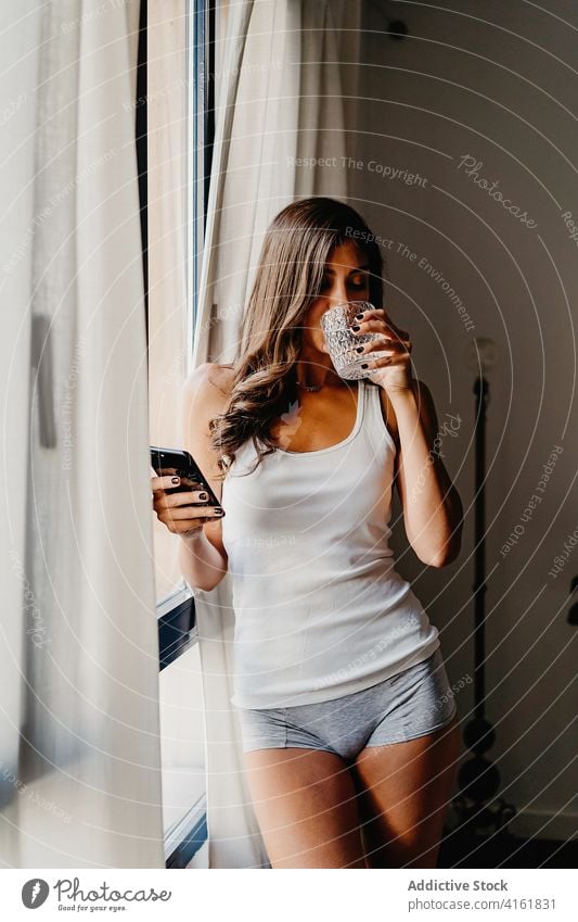 Junge Frau mit einem Glas Wasser und einem Smartphone am Fenster stehend am Morgen trinken benutzend heimwärts Browsen Apparatur tausendjährig Lifestyle