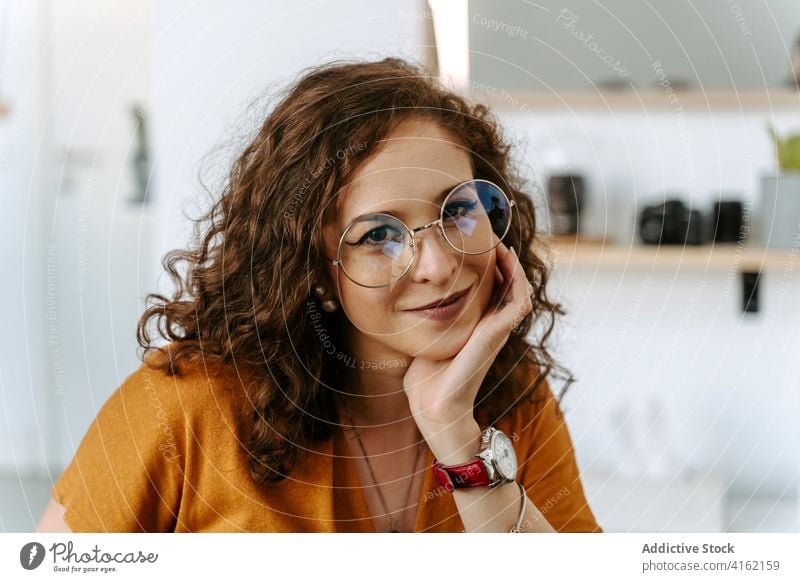 Glückliche junge Frau sitzt am Tisch mit der Hand am Kinn positiv Lächeln Stil heiter Unternehmer Inhalt froh Freude Ingwerhaar krause Haare lässig Brille