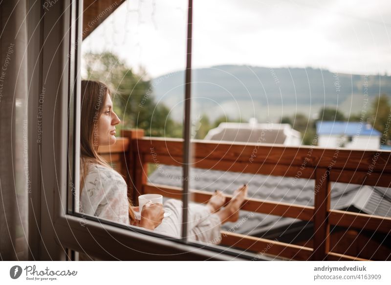 Junge Frau stehend nach dem Duschen am Morgen auf dem Balkon des Hotels. hält eine Tasse Kaffee oder Tee in ihren Händen. Blick nach draußen Natur Wald und Berg