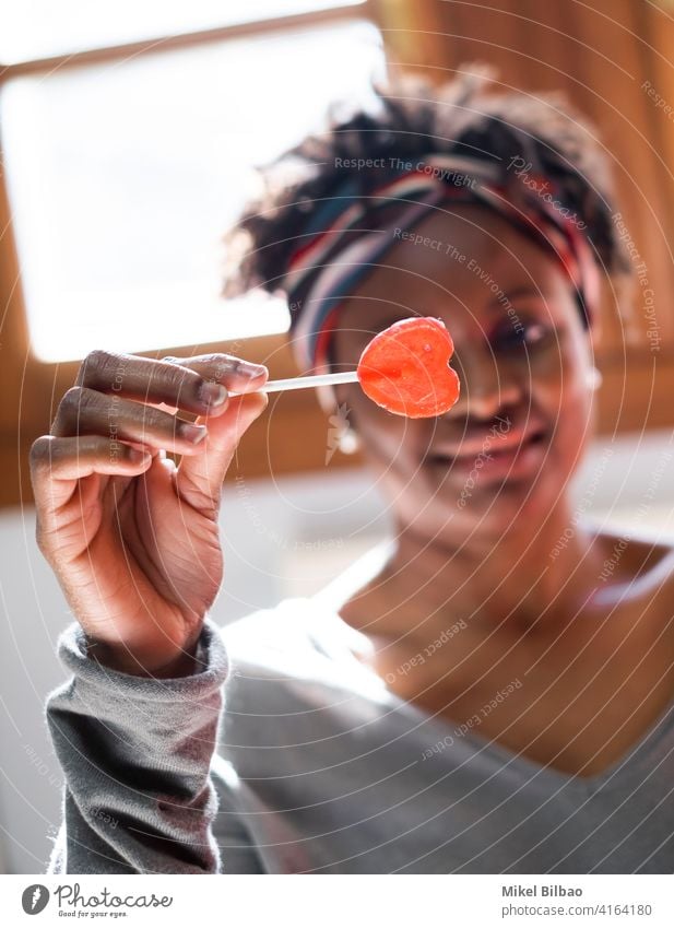 Junge schwarze Frau zu Hause lächelnd mit einem Lutscher in der Hand Schönheit Afroamerikaner Afrikanisch Menschen Dame Model Ausdruck Frisur im Innenbereich