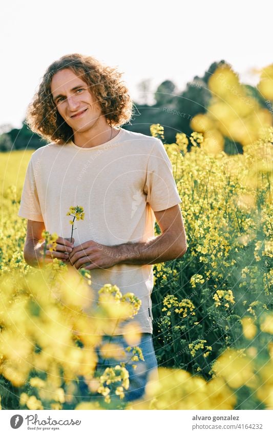 junger Mann mit lockigem Haar mit einer gelben Blume in einem Rapsblütenfeld Frühling Natur Landschaft che männlich Porträt Typ gutaussehend Person attraktiv