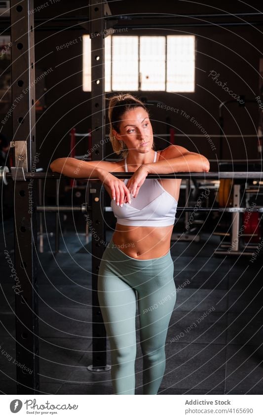 Entschlossene Sportlerin steht in der Nähe von Langhantel im Fitnessstudio Curl-Hantel ruhen Training muskulös stark Athlet Gerät selbstbewusst Frau BH Leggings