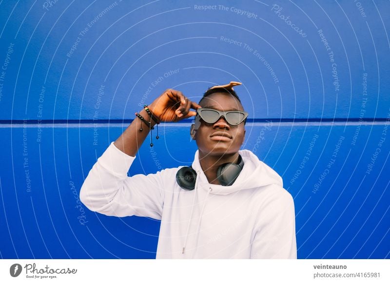 Junger afroamerikanischer schwarzer Mann mit weißem Sweatshirt, Sonnenbrille, Kamm und Kopfhörern an einer blauen Wand mit Vertrauen jung Afrikanisch Afro-Look