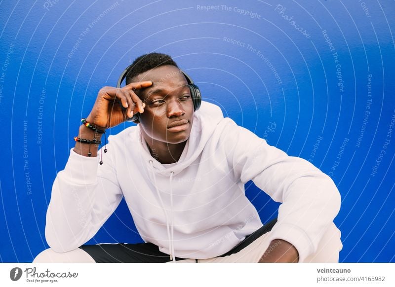 Junger afroamerikanischer schwarzer Mann mit weißem Sweatshirt und Kopfhörern, der an einer blauen Wand sitzt und zur Seite schaut. Moderner Look jung