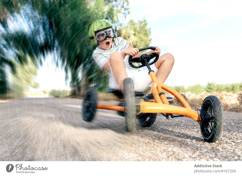 Aufgeregter Junge fährt Tretauto auf der Straße Mitfahrgelegenheit erstaunt Erholung Spaß haben Aktivität aufgeregt Schutzbrille Kindheit trendy anhaben Go-Kart