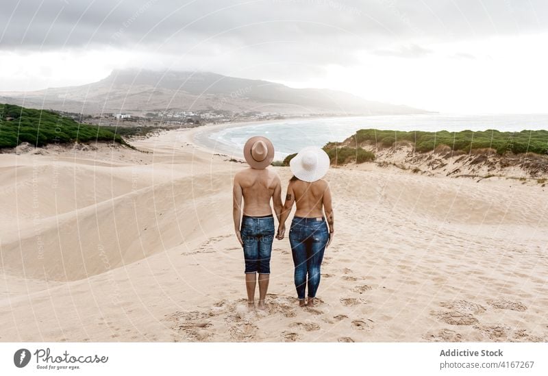 Unerkennbares Paar ohne Hemd, das sich an den Händen hält und an einer sandigen Küste steht oben ohne Meeresufer Strand erstaunlich ruhig Hafengebiet