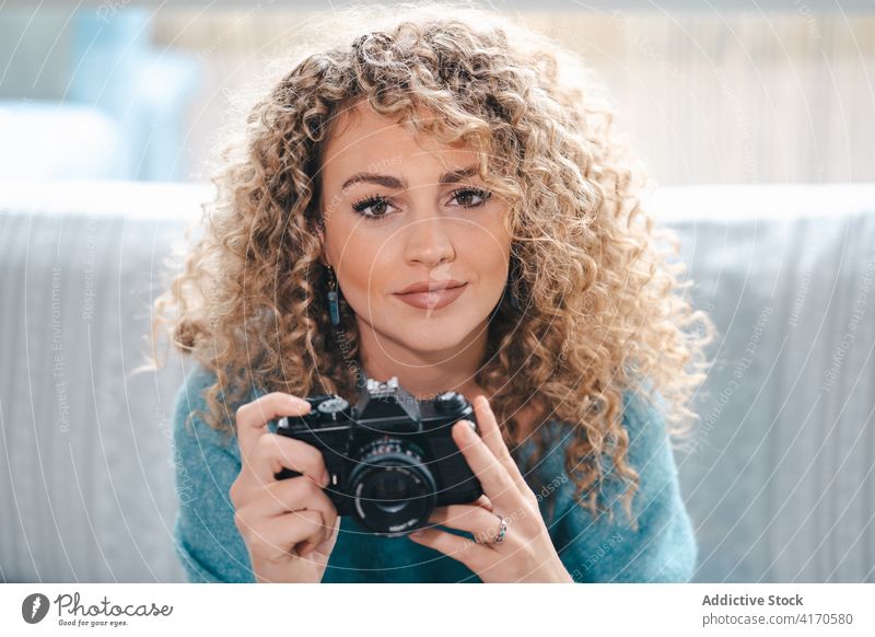 Fröhliche Frau mit Fotokamera zu Hause Fotograf fotografieren heiter heimwärts Talent gemütlich Fotoapparat Fotografie krause Haare hell Appartement lässig