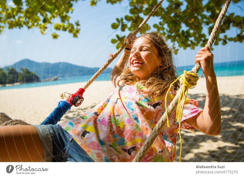 Glückliche niedliche kleine Mädchen lachen und schwingen auf Schaukel auf dem Baum am Strand. Schöne Sommer sonnigen Tag, türkisfarbenes Meer, Felsen, weißer Sand, malerische tropische Landschaft. Phuket, Thailand. Unbeschwert