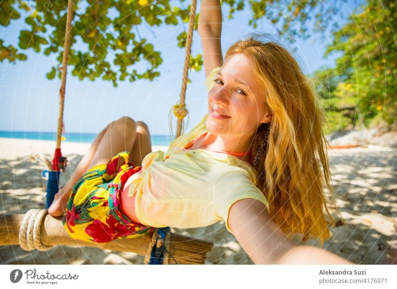 Glückliche junge Frau mit roten Haaren lachen und schwingen auf Schaukel auf einem Baum am Strand, ein Selfie zu nehmen. Schöne Sommer sonnigen Tag, türkisfarbenes Meer, weißer Sand, tropische Landschaft. Phuket, Thailand.