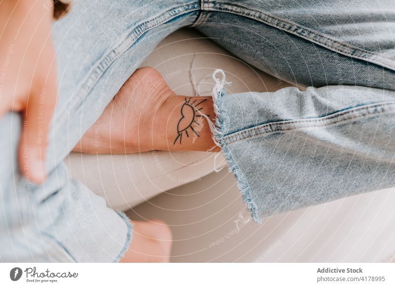 Crop-Frau mit Tattoo am Bein kreativ Sonne Form Kunst Design jung Individualität Kultur Mode cool Persönlichkeit sitzen Jeanshose Sofa ruhen Stil Dame