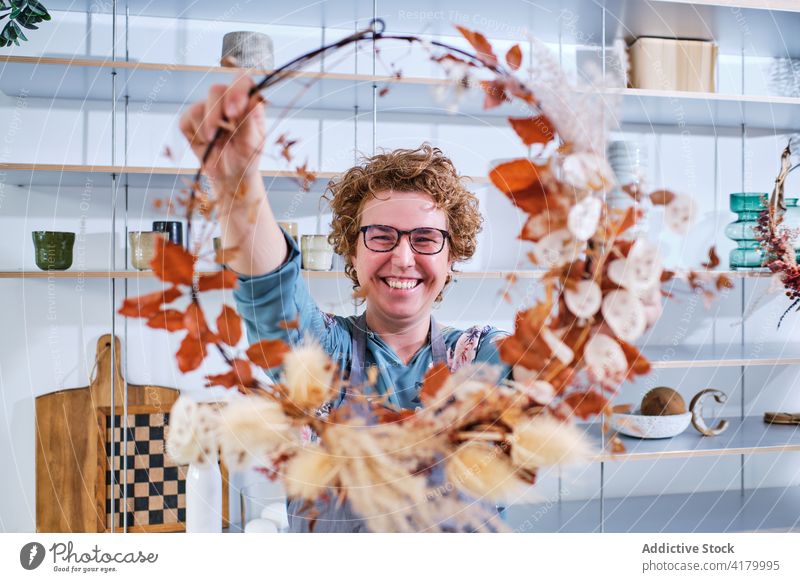 Fröhliche Frau mit handgefertigtem Blumenkranz im Geschäft Totenkranz Blumenhändler Basteln Werkstatt getrocknet Blatt Herbarium kreativ Salon Pflanze Lächeln