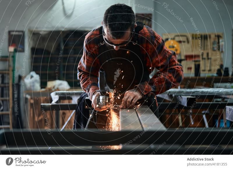 Unbekannter männlicher Schmied, der ein Eisenteil von Hand mit einer Maschine in einer Schmiede schleift Mann polnisch Schleifmaschine Metallbearbeitung