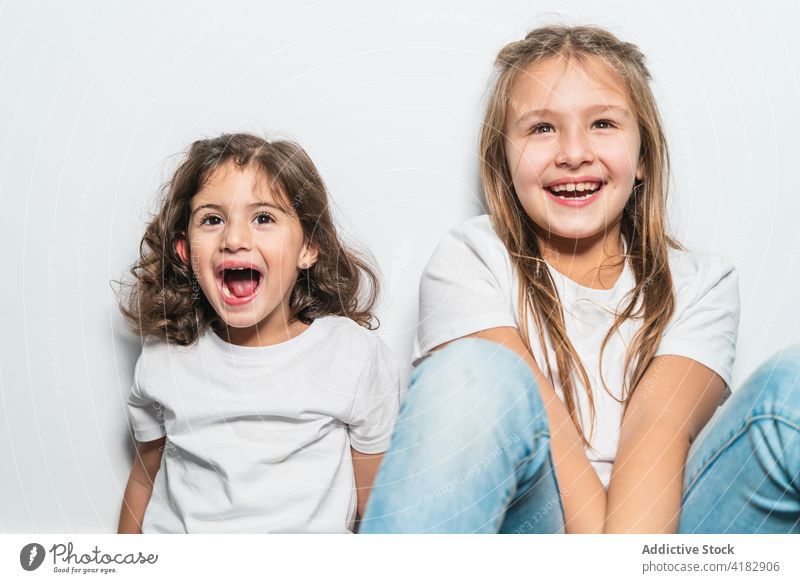 Fröhliche kleine Schwestern sitzen auf dem Boden Mädchen heiter Glück Stock niedlich Kindheit Zusammensein Freude zu Hause Zahnfarbenes Lächeln genießen