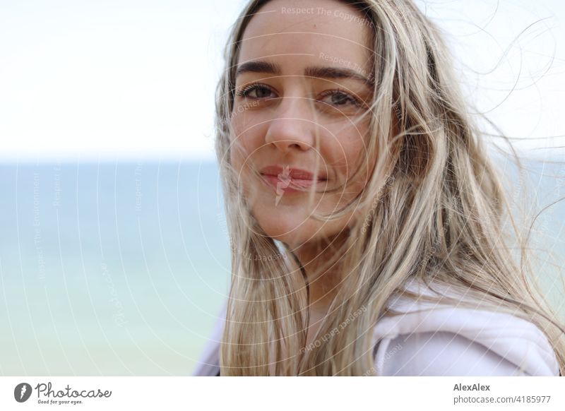 Ein schönes blondes Mädchen steht am Ostseestrand und schaut in die Kamera Lächeln freudig Landschaft von Freude Außenaufnahme Gesicht Junge Frau hübsch