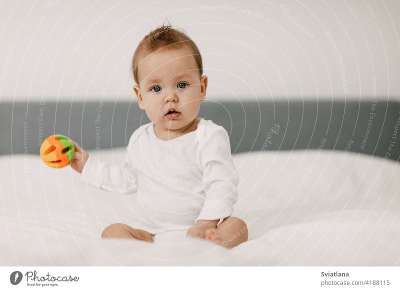 Das Kind sitzt in einem schneeweißen Bett, lacht und spielt mit einer Rassel. Baby Familie Spaß Sitzen Spielzeug Gesundheit heimwärts Schneewittchen