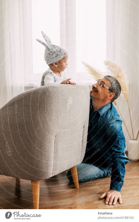 Kind in lustigen Osterhasen-Kostüm sitzt auf einem Stuhl, spielt mit Papa und lacht Baby Kaninchen Anzug Ostereier Spiel Sitzen Spielzeug Bett Innenbereich