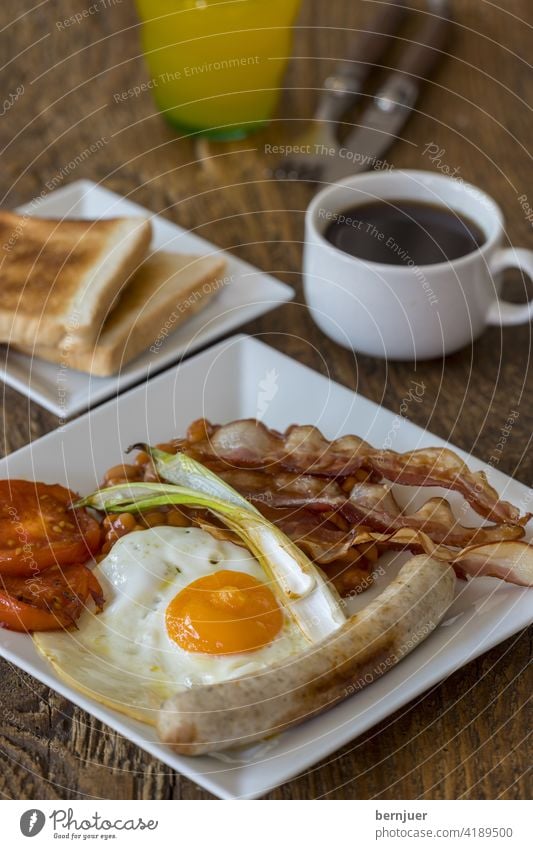 Nahaufnahme eines englischen Frühstücks Tomate Zwiebel Seite SpiegelEi Toast Speck rustikal bohne Fleisch Pilz Wurst Teller Butter Gericht traditionell morgen