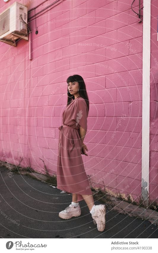 Modische junge ethnische Dame steht in der Nähe von rosa Wand und schaut weg Frau besinnlich nachdenklich Stil cool Mode Windstille Straße verträumt traumhaft