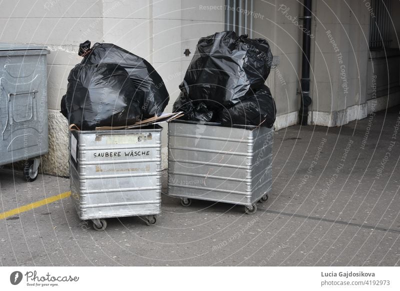 Waste management and two bed sheet carts or trolleys with big full black plastic bags in hotel backyard. On one container there is inscription in German language saying clean sheets.