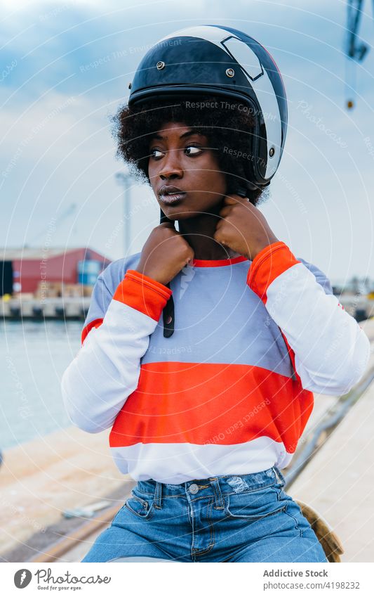 Seriöse junge ethnische Frau mit Helm auf dem Motorrad sitzend Seeküste Vorschein Persönlichkeit trendy Stauanlage allein Biker cool reisen Fahrzeug schwarz