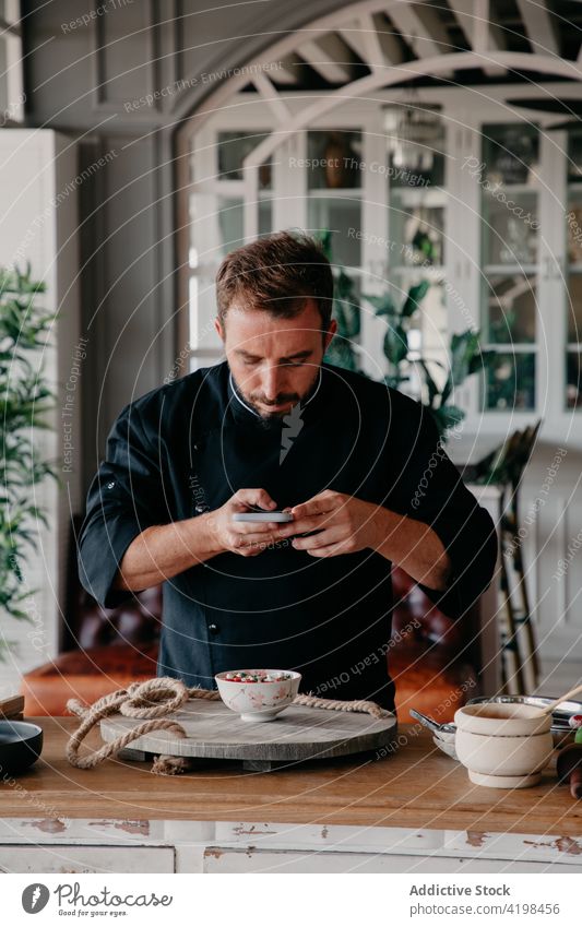 Seriöser Koch verwendet Smartphone zum Fotografieren von Essen in der Schüssel Mann Küchenchef benutzend fotografieren kulinarisch Mahlzeit Restaurant schießen