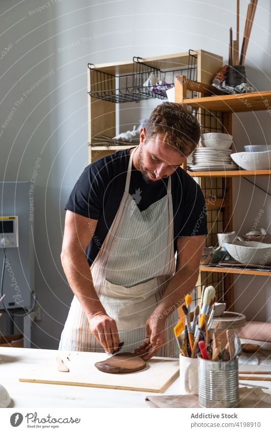 Bärtiger junger Mann bei der Herstellung einer Tonplatte während der Arbeit im Atelier kreieren Tonwaren Werkstatt Lächeln kneten Meister Töpferwaren positiv