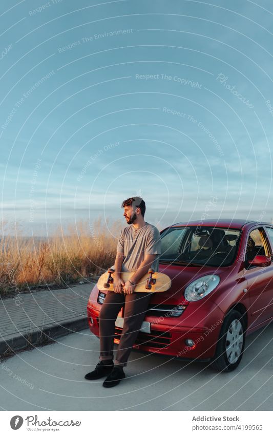 Junger bärtiger Skater, der sich auf die Motorhaube eines Autos stützt und in die Natur schaut Mann anlehnen PKW Freiheit Landschaft ländlich abgelegen Straße