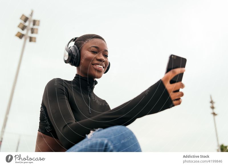 Lächelnde schwarze Frau mit Headset beobachtet Smartphone auf der Straße zuschauend Afroamerikaner Gesang zuhören Kurze Haare Inhalt Internet Porträt benutzend