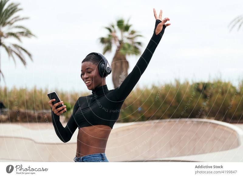 Lächelnde schwarze Frau mit Headset beobachtet Smartphone auf der Straße zuschauend Afroamerikaner Gesang zuhören Kurze Haare Inhalt Internet Porträt benutzend