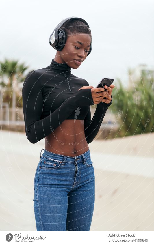 Lächelnde schwarze Frau mit Headset beobachtet Smartphone auf der Straße zuschauend Afroamerikaner Gesang zuhören Kurze Haare Inhalt Internet Porträt benutzend