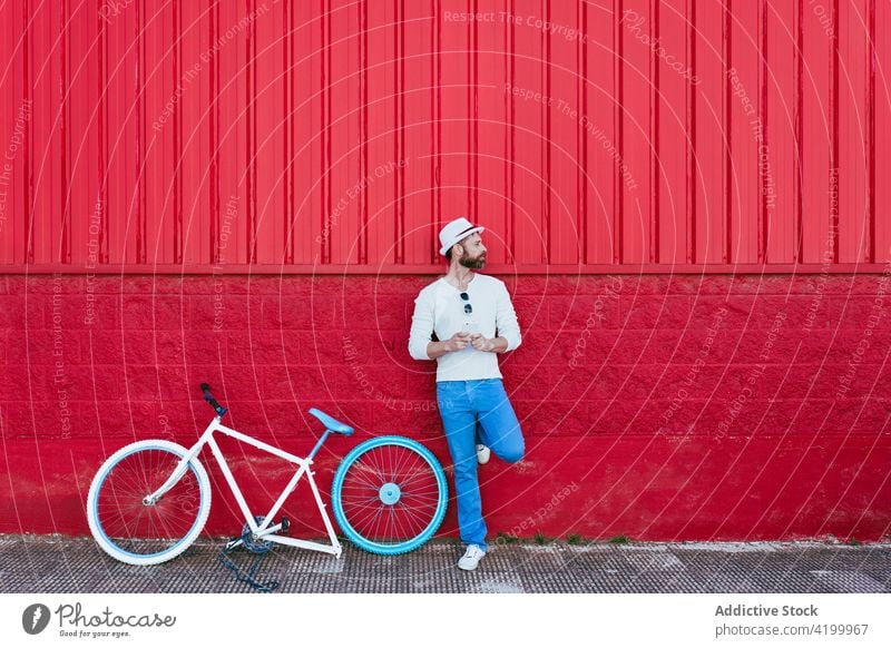 Mann mit Smartphone steht in der Nähe von Fahrrad gegen rotes Gebäude sich[Akk] entspannen Straße trendy Stil rote Wand ruhen Mode nachdenklich Großstadt sonnig