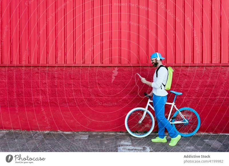 Mann mit Smartphone und Kopfhörern geht mit Fahrrad an rotem Gebäude vorbei Straße trendy Stil rote Wand ruhen Mode Großstadt urban männlich Vollbart cool