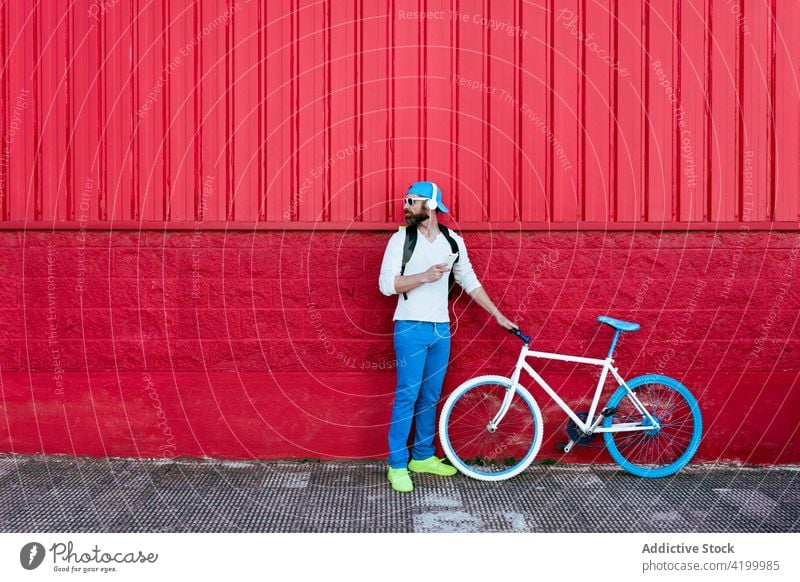 Mann mit Smartphone und Kopfhörern geht mit Fahrrad an rotem Gebäude vorbei Straße trendy Stil rote Wand ruhen Mode Großstadt urban männlich Vollbart cool