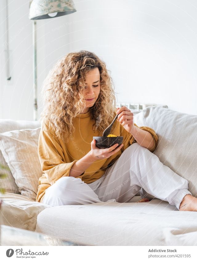 Blonde Frau sitzt mit einer Schüssel mit gesundem Essen auf dem Sofa positiv Appartement Lebensmittel Schalen & Schüsseln krause Haare Mango Gesundheit jung