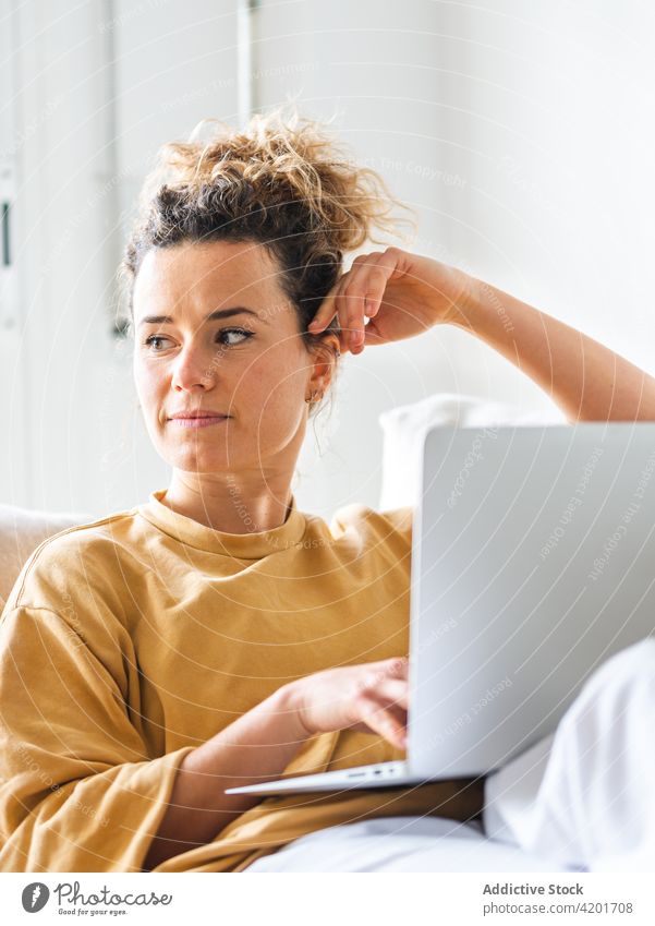 Lächelnde Frau sitzt mit Laptop auf dem Sofa in einem hellen Raum benutzend Browsen Appartement heimwärts Gerät freiberuflich nachdenklich positiv lässig